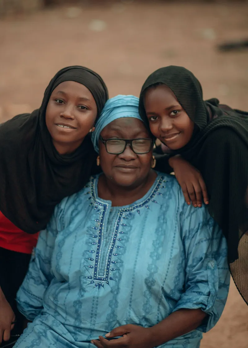 An older woman in a blue dress and glasses, smiling with two young women in hijabs